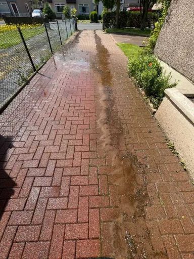 Wet pathway with a herringbone pattern and water flow, bordered by grass and a fence.