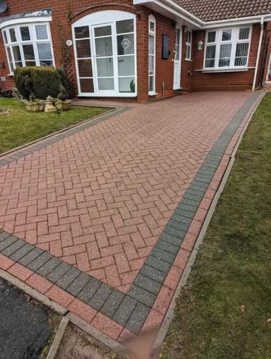 Paved driveway with red and grey blocks alongside a lawn and brick house.